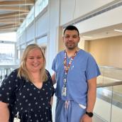 A woman stands with a man, facing the camera. She has long blonde hair, a black and white shirt and he is a doctor with medical scrubs, blue, and a lanyard.