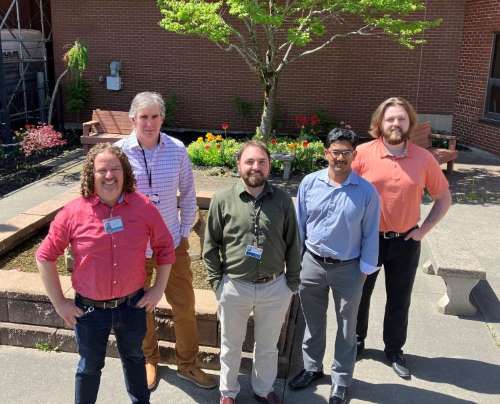 Five men standing in front of a garden. All have their hands on their sides, looking up to a camera. From left to right, one man with long curly blond hair wearing a red shirt and jeans, man behind him has grey hair with a plaid button up and brown khakis, third man has brown hair and beard wearing a green button up and beige pants, fourth man has black hair wearing a blue button up and grey dress pants, the last man has long blond hair with brown beard wearing a peach coloured shirt and black pants.