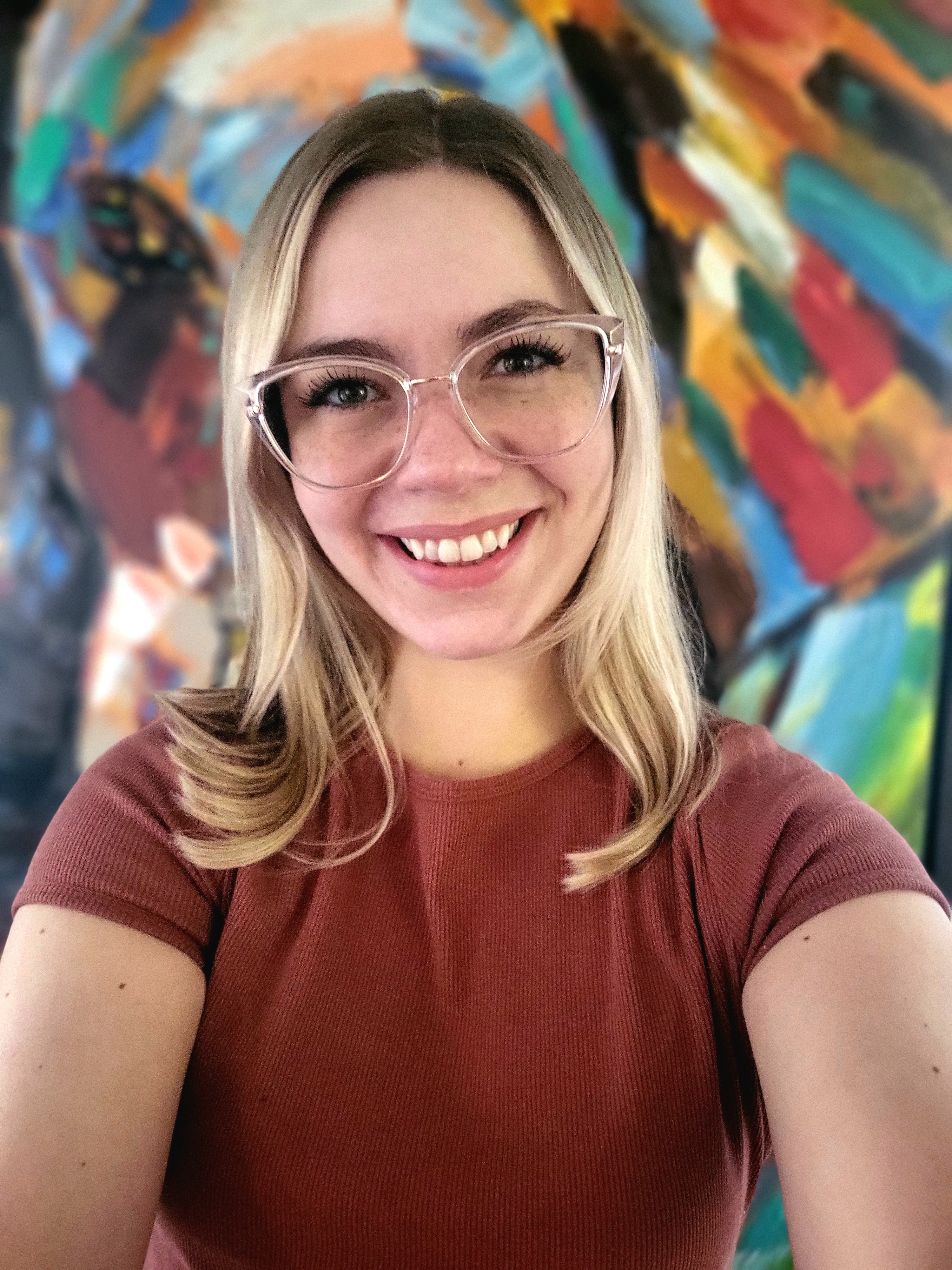 Woman in front of a colourful painting. She has blonde hair and is wearing a brown shirt and clear framed glasses.
