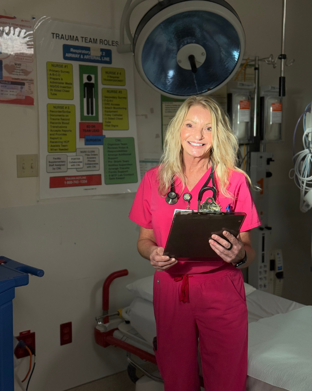 Woman standing in front of a hospital bed, with blonde long hair, wearing pink scrubs, holding a clipboard and has a stethoscope on her neck.