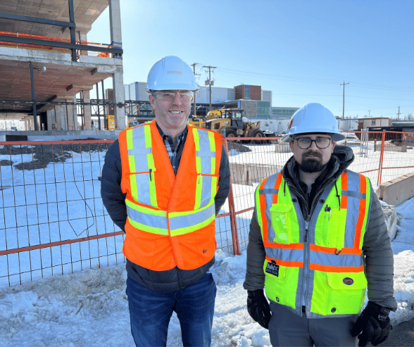 Two men stand together wearing safety vests, hardhats and glasses at a construction site and are smiling at the camera.