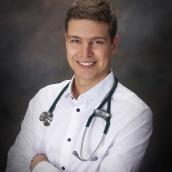A man with short brown hair is wearing a white doctor's coat and stethoscope is standing and smiling with his arms crossed looking at the camera.