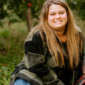 A woman is sitting outdoors on a blanket, smiling at the camera. She has long hair and is wearing a fall jacket with jeans.