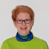 Woman with short, red hair and glasses with black frames is wearing a sweater and is smiling at the camera.