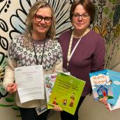 Two adult females standing facing camera holding educational literature.