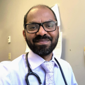 Dr. Nag is in his office, he wears a white dress shirt with a purple-printed tie and has a stethoscope around his neck.