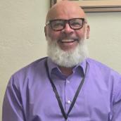 Man with bald head and white beard, wearing glasses, smiles at camera.