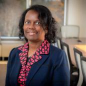 A black woman with chin length dark hair, wearing a blazer, smiles at camera; boardroom in background.