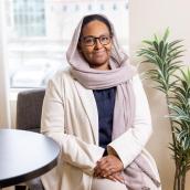 Photo of woman smiling, wearing beige head scarf and glasses, navy top, long off-white blazer, beige linen pants.