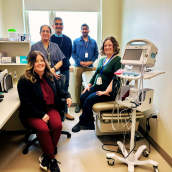 A group of five smiling healthcare providers—two nurse practitioners and three physicians—pose together in the Colchester Cardiac Clinic. One is sitting on a hospital bed, while others stand beside them in the clinic space.