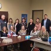 Bariatric care teams from Truro and Glace Bay are gathered around a table in a conference room wearing casual, professional clothes. Seven people are sitting and nine others are standing behind them.