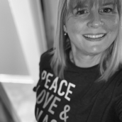 Woman with shoulder-length, straight hair and bangs smiles at the camera. She is wearing a black t-shirt with the words "Peace Love & Vaccine" in white and grey letters across the front.