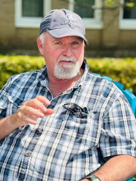 A man is sitting wearing a green and yellow plaid shirt, light blue ball cap and he has a white goatee.