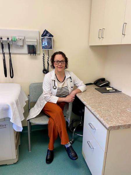 Woman sits in a chair at a desk in a doctor's office. She has short brown, curly hair and is wearing a white lab coat, a black and white striped shirt and red pants with black shoes.
