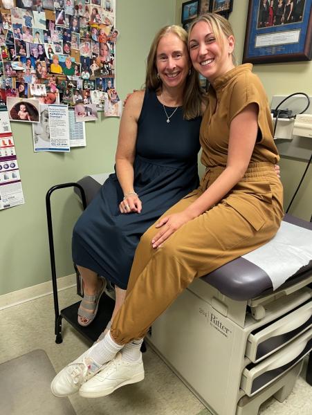 Photo Dr. Kathy Gallagher and Dr. Allison Verge in their clinic sitting on a patient exam table, smiling at the camera