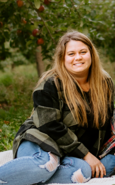 A woman is sitting outdoors on a blanket, smiling at the camera. She has long hair and is wearing a fall jacket with jeans.