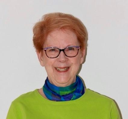 Woman with short, red hair and glasses with black frames is wearing a sweater and is smiling at the camera.