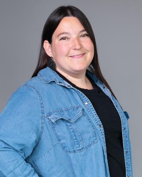 A woman with long hair smiles at the camera, wearing a button-up shirt over a t-shirt. She has beaded earrings, and the background is neutral.