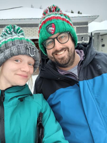 Two people wearing winter hats and jackets are standing together outdoors.