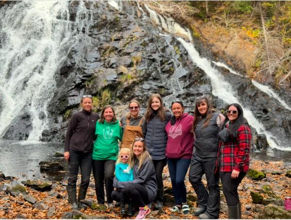 A group of nine people, pose in front of a cascading waterfall in an outdoor setting during autumn. They are dressed in warm clothing, including jackets, hoodies, and boots, with some wearing sunglasses.