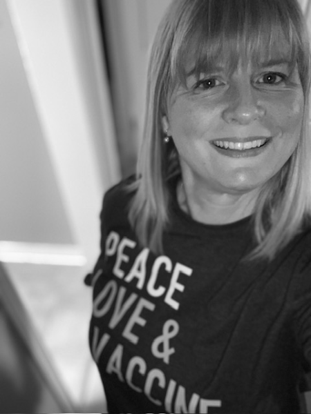 Woman with shoulder-length, straight hair and bangs smiles at the camera. She is wearing a black t-shirt with the words "Peace Love & Vaccine" in white and grey letters across the front.